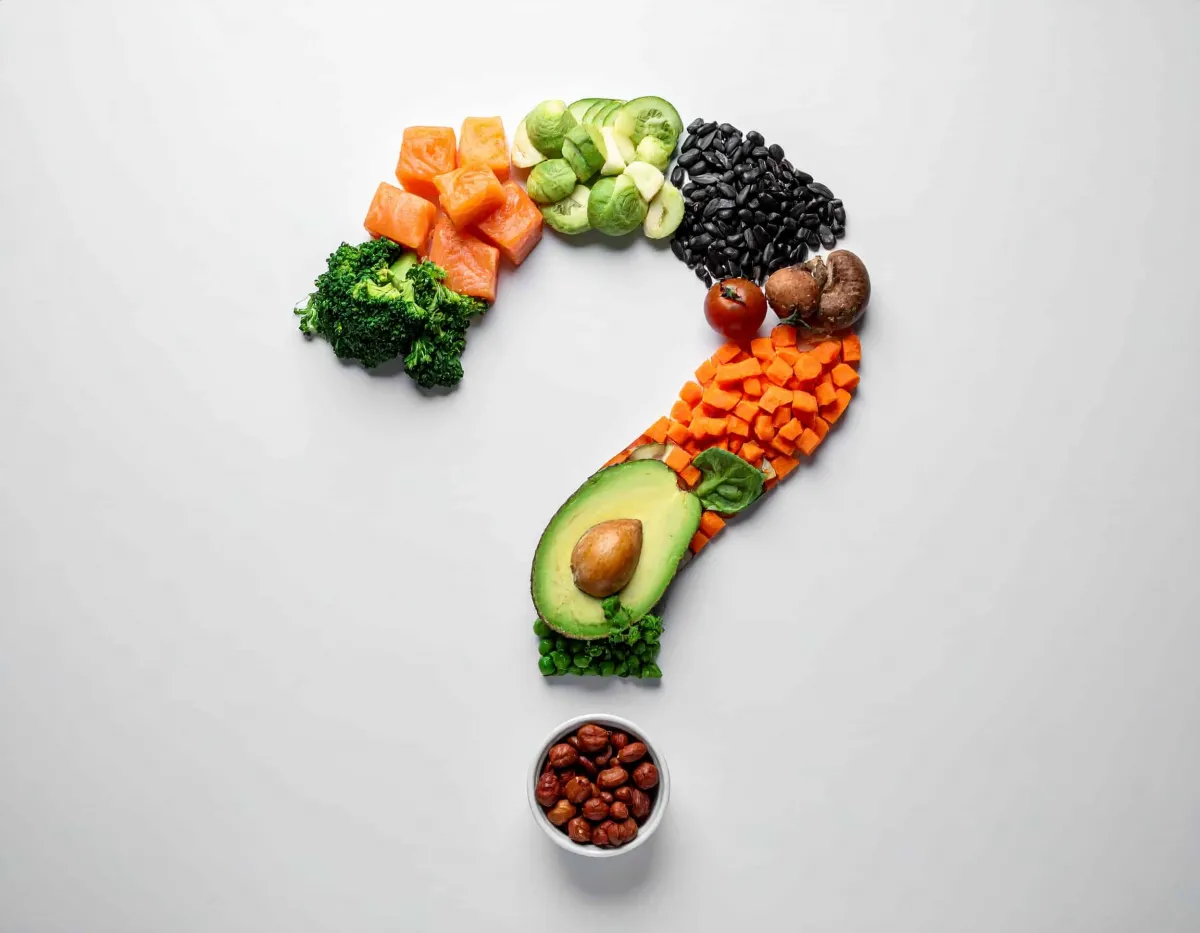 Verschiedene frische Lebensmittel sind auf einem weißen Hintergrund kreativ zu einem Fragezeichen arrangiert. Der Bogen besteht aus Lachs, Brokkoli, Rosenkohl, Gurke, schwarzen Bohnen, Champignon, Tomate, Karotten, Avocado und Erbsen. Den Punkt des Fragezeichens bildet eine kleine Schale mit Haselnüssen. Das Bild symbolisiert die Frage nach gesunder Ernährung.