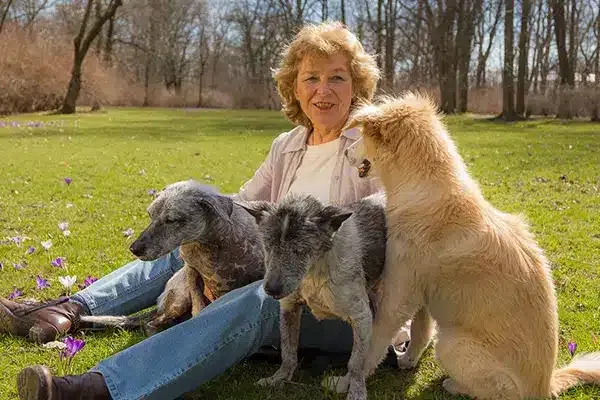Eine lächelnde ältere Frau mit kurzen, lockigen, rotblonden Haaren und hellrosa Jacke sitzt auf einer Wiese im Park und ist von drei Hunden umgeben: einem grauen Mischlingshund, einem grau-weißen Mischlingshund und einem goldbeigen, größeren Hund. Rund um sie blühen lila Krokusse, im Hintergrund sind kahle Frühlingsbäume zu sehen.