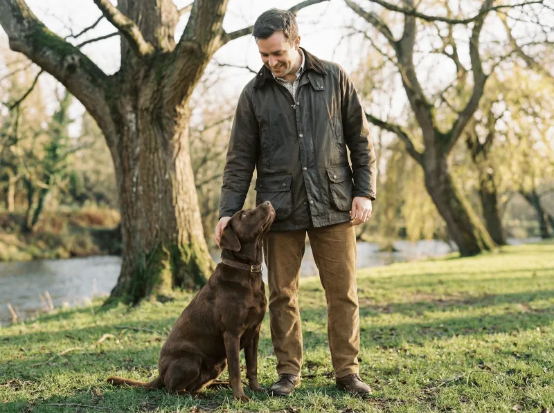 Ein lächelnder Mann mittleren Alters in einer olivgrünen Wachsjacke und brauner Cordhose steht auf einer Wiese und schaut auf einen sitzenden Chocolate Labrador mit braunem Lederhalsbandherunter. Der Hund blickt aufmerksam zu seinem Besitzer auf, während dieser ihn sanft am Kinn berührt. Im Hintergrund kahle Bäume, ein Fluss und weiches Herbst- bzw. Winterlicht.