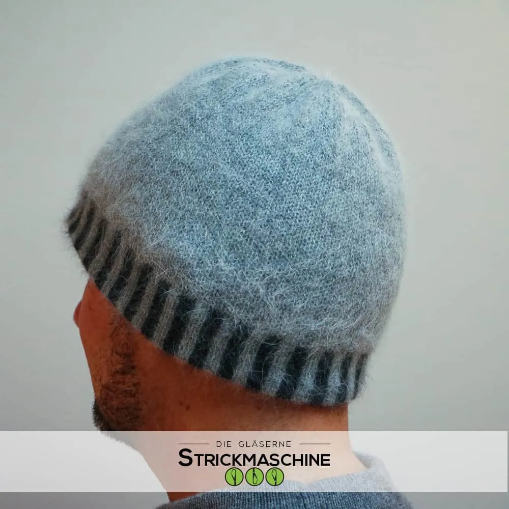 Rückenansicht eines Mannes mit Bart, der eine selbst gestrickte Beanie-Mütze aus flauschigem Angoragarn in hellem Eisblau mit dunkelgrau-gestreiftem Rippenbündchen trägt. Die Mütze hat eine weiche, zarte Textur. Darunter ein grauer Pullover. Vor neutralem hellgrauem Hintergrund. Unten das Logo von „Die Gläserne Strickmaschine".