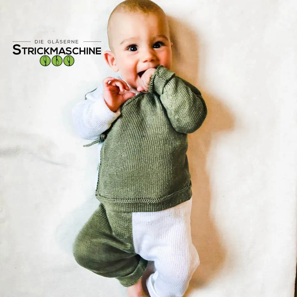 Ein fröhlich lächelndes Baby liegt auf dem Rücken und trägt ein selbst gestricktes zweiteiliges Set in Olivgrün und Weiß — bestehend aus einem Pullover mit weißen Ärmelbündchen und einer olivgrünen Hose mit weißem Einsatz. Das Baby steckt die Hand in den Mund und schaut aufgeweckt in die Kamera. Oben links das Logo von „Die Gläserne Strickmaschine".