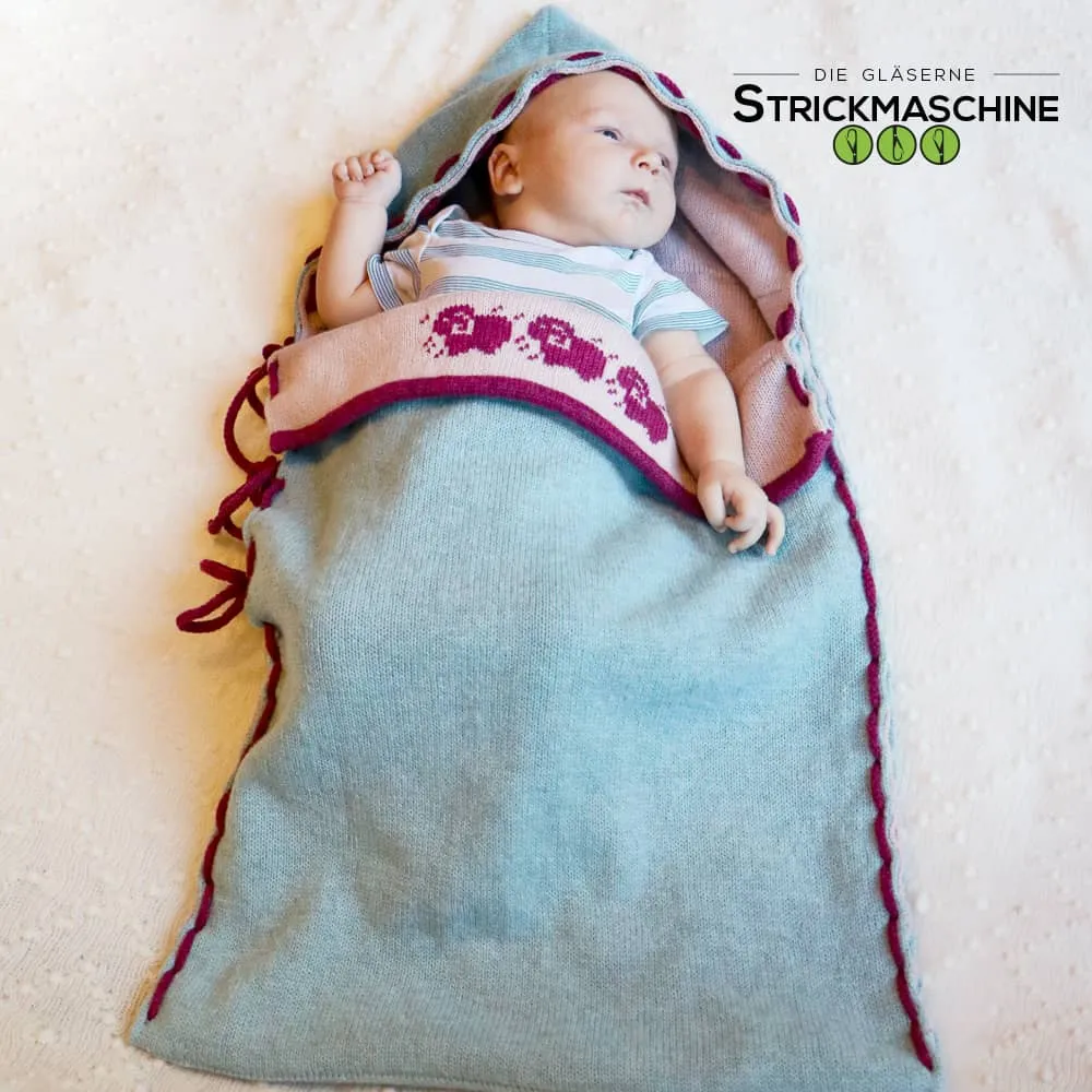 Ein Neugeborenes liegt wach und entspannt in einem selbst gestrickten Babyschlafsack in Hellblau mit bordeauxroten Zierkanten und Schnüren. Der Schlafsack hat eine Kapuze mit Wellenkante und einen rosafarbenen Innenkragen mit gestricktem Schaf-Jacquardmuster in Bordeaux. Das Baby trägt ein gestreiftes Kurzarm-Body. Oben rechts das Logo von „Die Gläserne Strickmaschine".