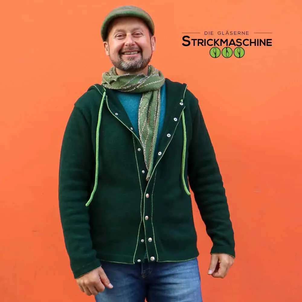 Ein lächelnder Mann mit Bart und olivgrünem Schiebermütze steht vor einer leuchtend orangen Wand und präsentiert einen selbst gestrickten dunkelgrünen Hoodie mit Druckknopfleiste und neongrünen Kordeln. Darunter trägt er ein türkises Shirt und einen gemusterten Strickschal in Grün- und Beigetönen sowie eine Jeans. Oben rechts das Logo von „Die Gläserne Strickmaschine".