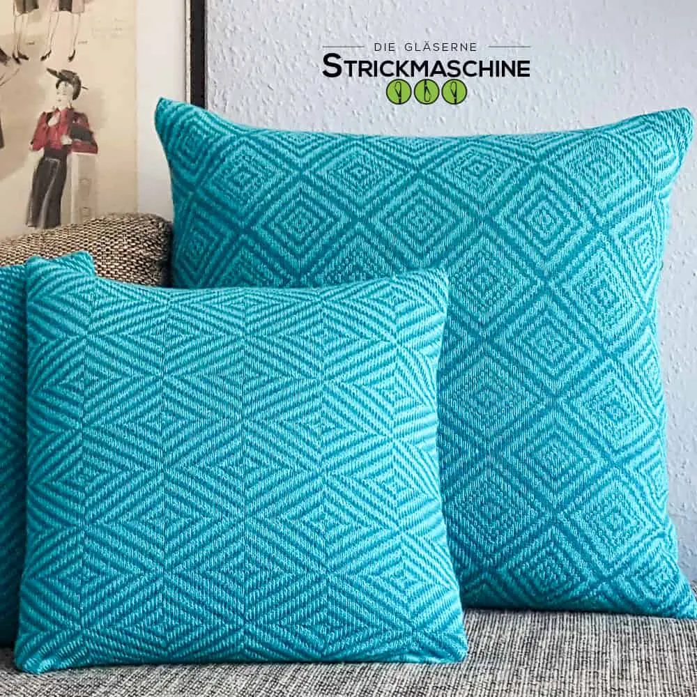 Drei selbst gestrickte Kissen in leuchtendem Türkisblau mit geometrischem Rautenmuster liegen auf einem grauen Sofa. Die Kissen sind in zwei verschiedenen Größen zu sehen und zeigen ein gleichmäßiges, strukturiertes Jacquard-Strickmuster. Im Hintergrund eine weiße Wand und ein gerahmtes Kunstposter. Oben das Logo von „Die Gläserne Strickmaschine".