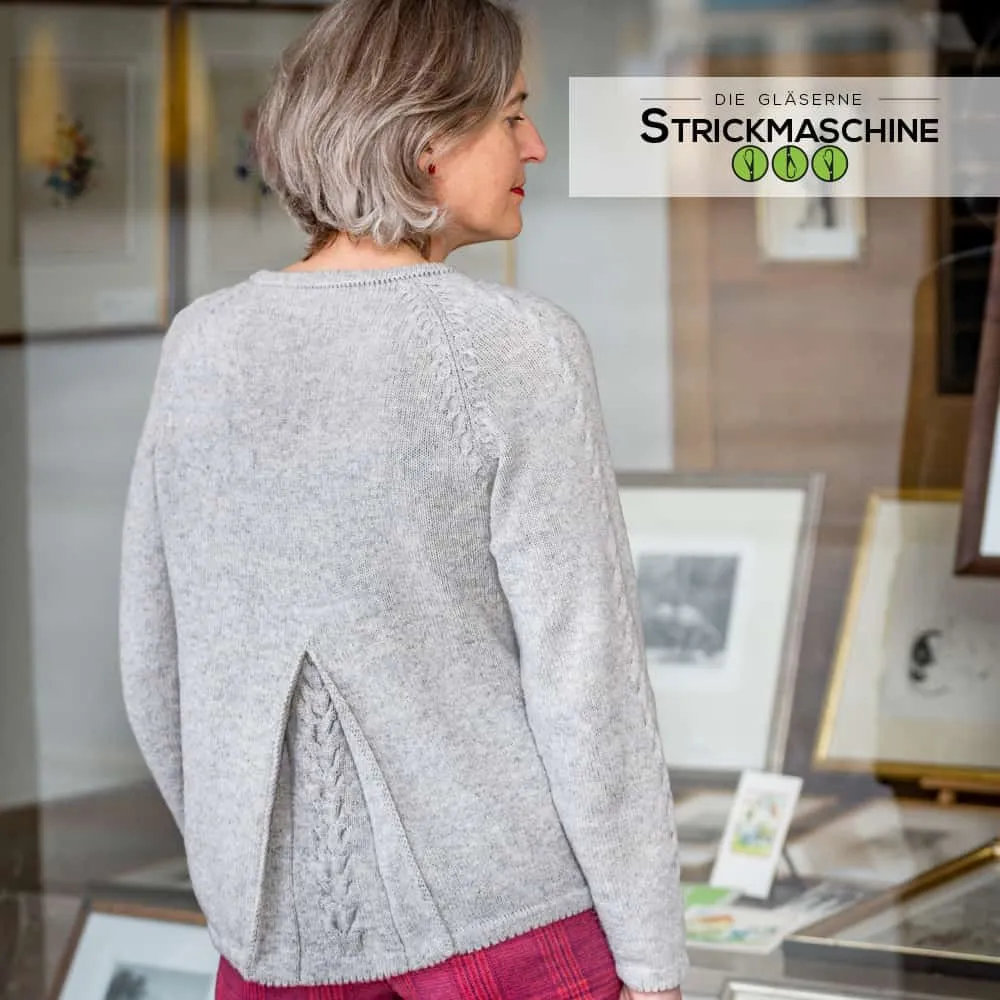 Rückenansicht einer Frau mit silbergrauem Bob, die einen selbst gestrickten hellgrauen Raglan-Pullover mit Zopfmuster-Details an den Schulternähten und einem dekorativen Schlitz mit Zopfmuster am unteren Rücken trägt. Dazu ein karierter Hosenrock in Pink-Rot. Im Hintergrund ein Innenraum mit gerahmten Kunstwerken. Oben rechts das Logo von „Die Gläserne Strickmaschine".