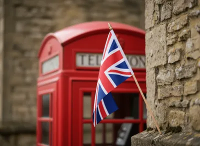Eine kleine Union-Jack-Flagge auf einem Holzstab lehnt an einer Natursteinmauer, im unscharfen Hintergrund eine klassische rote britische Telefonzelle mit der Aufschrift „TELEPHONE".