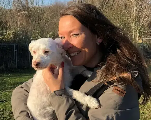 Lächelnde Frau mit langen braunen Haaren und grauer Jacke hält einen kleinen weißen Lockenhund (vermutlich Pudelmix) an ihre Wange gedrückt. Im Hintergrund eine grüne Außenanlage mit Bäumen und Zaun im Gegenlicht.