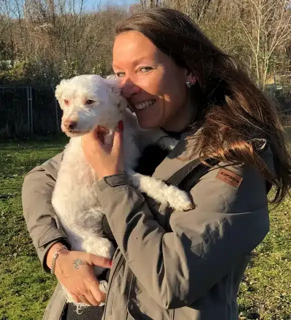 Lächelnde Frau mit langen braunen Haaren und grauer Jacke hält einen kleinen weißen Lockenhund (vermutlich Pudelmix) an ihre Wange gedrückt. Im Hintergrund eine grüne Außenanlage mit Bäumen und Zaun im Gegenlicht.