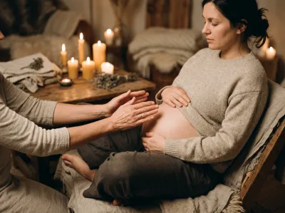 Schwangere Frau im beigen Strickpullover sitzt mit geschlossenen Augen entspannt auf einem Stuhl, während eine zweite Person sanft ihre Hände auf den freigelegten Babybauch legt. Im Hintergrund ein warmtoniges Zimmer mit mehreren brennenden Kerzen und getrockneten Kräutern auf einem Holztisch.