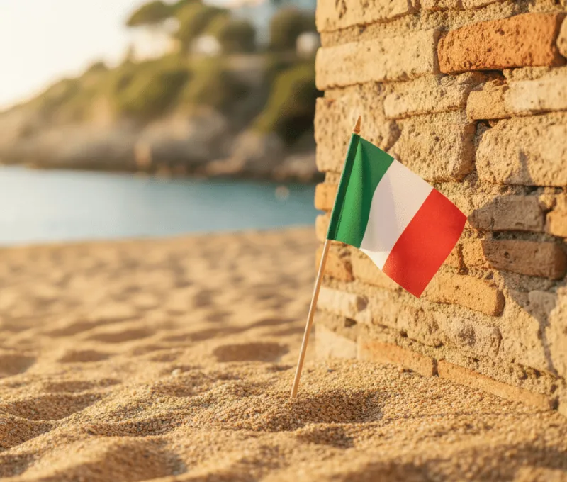 Eine kleine italienische Flagge steckt im Sand neben einer alten Steinmauer, im Hintergrund sind Strand, Meer und Küste unscharf zu sehen.