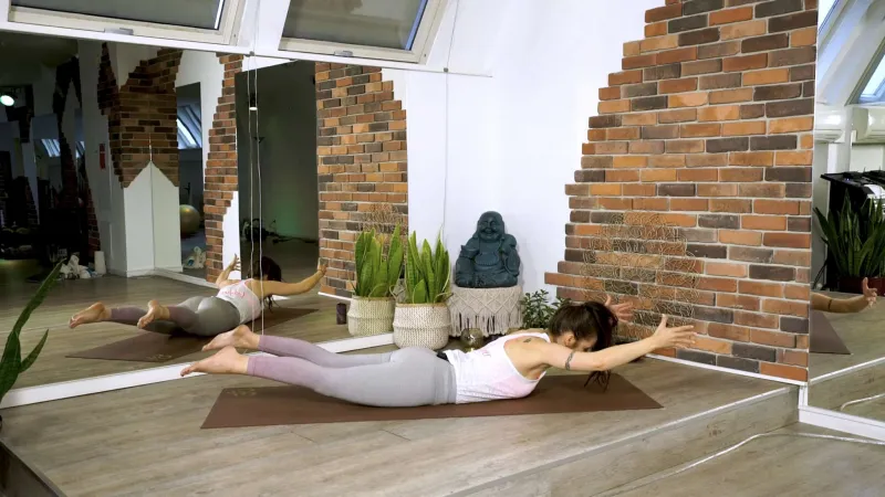 Screenshot eines Pilates-Onlinekurses: Eine Trainerin in weißem Top und hellgrauen Leggings führt die Pilates-Übung „Swimming