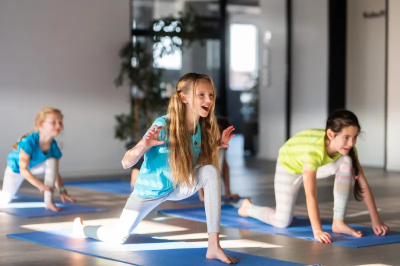 Kinder machen gemeinsam Yogaübungen auf blauen Matten in einem hellen Raum und haben dabei sichtbar Spaß.
