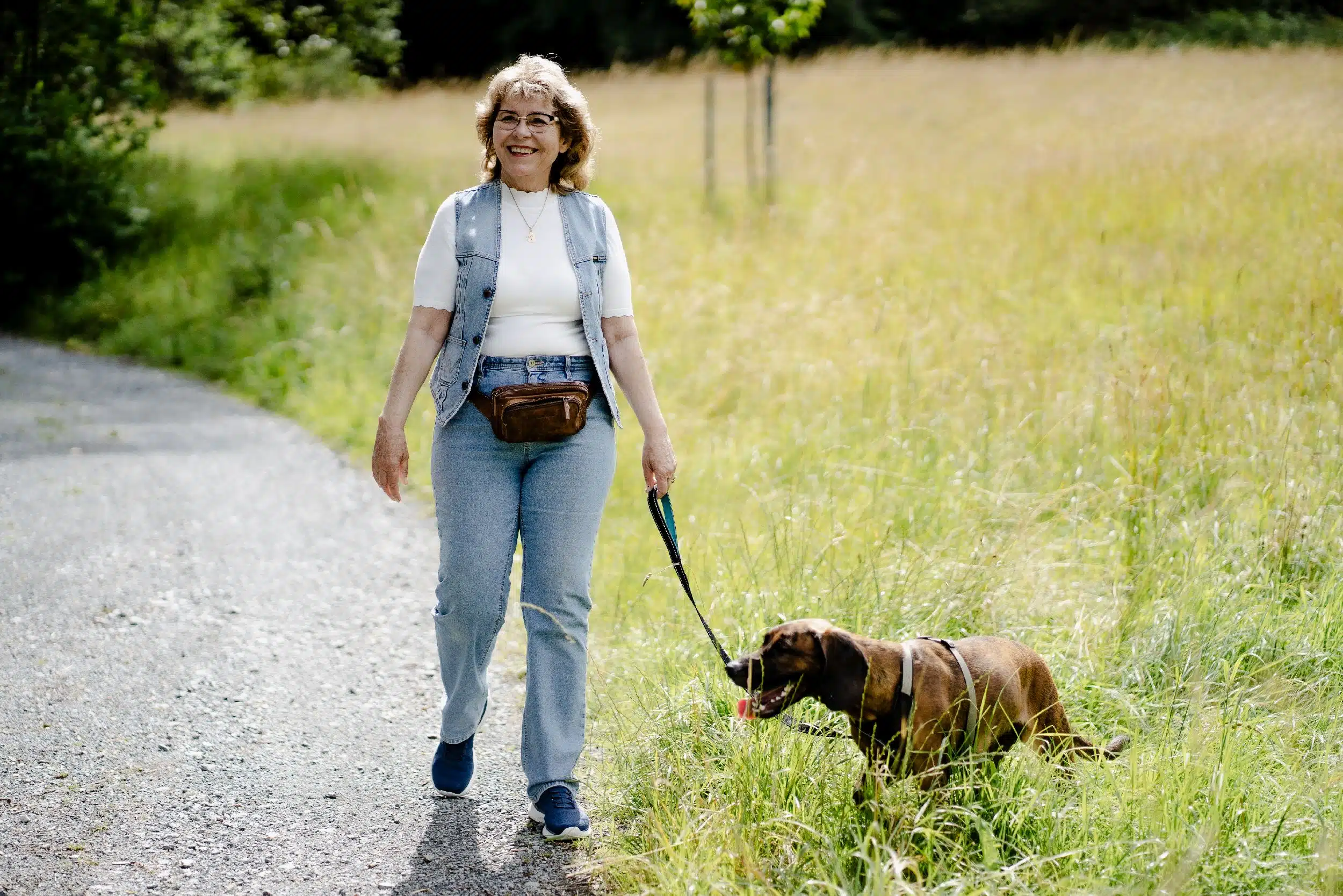 Eine fröhlich lächelnde Frau mittleren Alters mit kurzen, lockigen, blonden Haaren und Brille geht einen Schotterweg entlang. Sie trägt ein weißes T-Shirt, eine Jeansweste, hellblaue Jeans, blaue Sneaker und eine braune Gürteltasche. An ihrer Seite läuft ein brauner Hund mit Geschirr an der Leine, der im hohen Gras am Wegrand schnüffelt. Im Hintergrund eine sommerliche Wiese und Bäume.