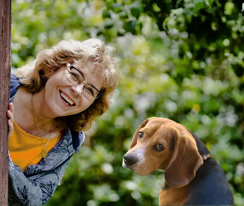 Eine herzlich lachende Frau mit kurzen, lockigen, blonden Haaren und Brille lehnt sich von links um einen Baumstamm und schaut einen Beagle mit schwarz-braunem Fell an. Der Hund sitzt ruhig daneben und blickt aufmerksam nach vorne. Im Hintergrund üppiges grünes Laub im Sonnenlicht.