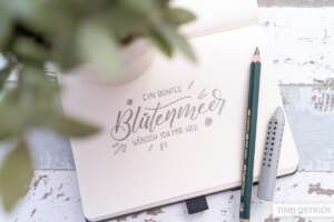 Aufgeschlagenes Bullet-Journal mit Bleistift-Lettering „Ein buntes Blütenmeer wünsch ich mir her" mit kleinen Blüten- und Blattdekors. Daneben ein Faber-Castell 9000 Jumbo-Bleistift und ein grauer Radiergummi-Aufsatz auf weißem Vintage-Holztisch. Foto: Timo Ostrich.
