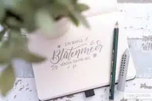 bluetenmeer - Webinare und Kurse Aufgeschlagenes Bullet-Journal mit Bleistift-Lettering „Ein buntes Blütenmeer wünsch ich mir her" mit kleinen Blüten- und Blattdekors. Daneben ein Faber-Castell 9000 Jumbo-Bleistift und ein grauer Radiergummi-Aufsatz auf weißem Vintage-Holztisch. Foto: Timo Ostrich.