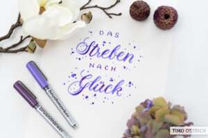 Handlettering „Das Streben nach Glück" in Lila-Violett-Brushschrift mit Farbspritzern auf weißem Papier. Daneben zwei Brushmarker Pro in Lila und Violett, eine weiße Magnolie, getrocknete Zapfen und eine Hortensie. Foto: Timo Ostrich.