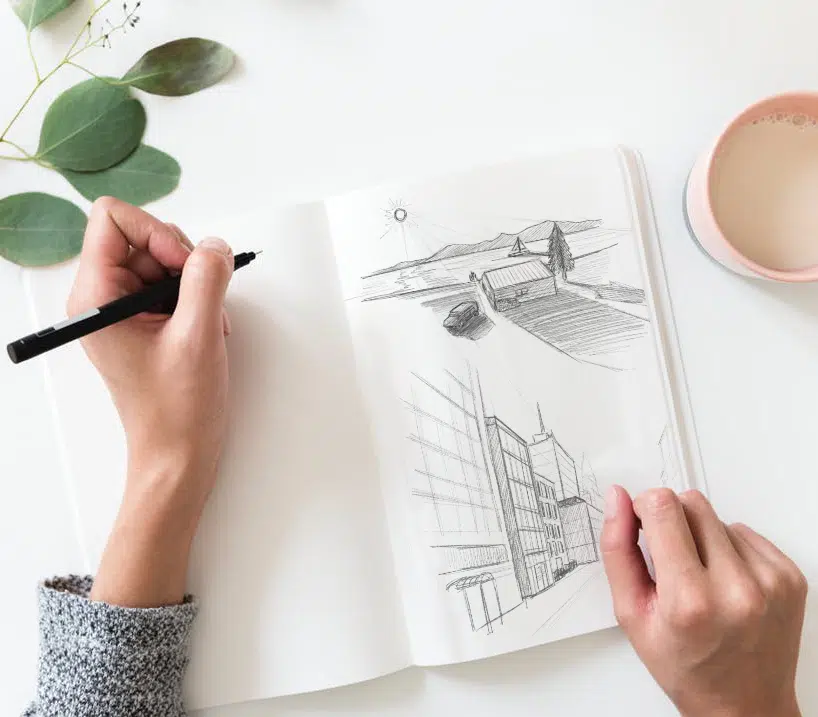 Draufsicht auf zwei Hände, die ein aufgeschlagenes Skizzenbuch halten und mit einem schwarzen Fineliner zeichnen. Im Skizzenbuch sind zwei Bleistiftzeichnungen zu sehen: eine Landschaft mit Haus, Baum und Bergen, sowie ein perspektivisches Stadtgebäude. Daneben auf dem weißen Tisch eine rosafarbene Kaffeetasse und ein Eukalyptuszweig.