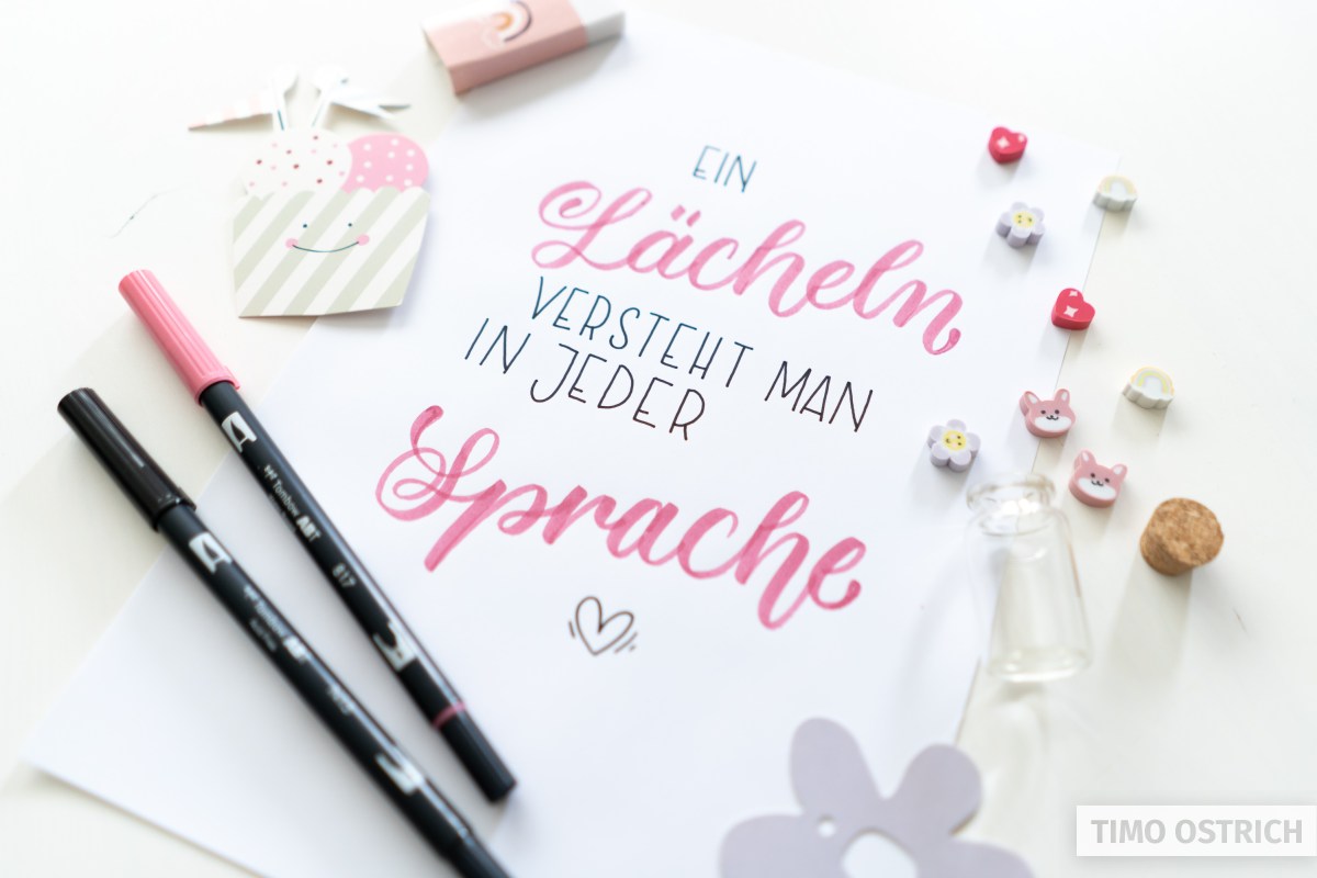 Handlettering „Ein Lächeln versteht man in jeder Sprache" in Pink und Petrol mit kleinem Herz auf weißem Papier. Daneben zwei Tombow ABT Dual Brush Pens, ein Herz-Anhänger, kleine Tier-Buttons, Glasfläschchen und dekorative Papierstanzen. Foto: Timo Ostrich.
