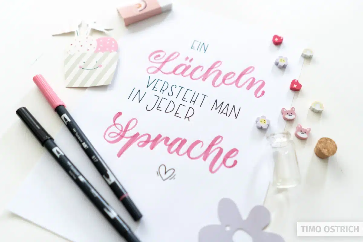 Handlettering „Ein Lächeln versteht man in jeder Sprache" in Pink und Petrol mit kleinem Herz auf weißem Papier. Daneben zwei Tombow ABT Dual Brush Pens, ein Herz-Anhänger, kleine Tier-Buttons, Glasfläschchen und dekorative Papierstanzen. Foto: Timo Ostrich.