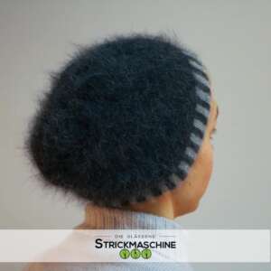 Eine Frau mit kurzem, grau-blondem Haar trägt einen selbst gestrickten großen Dreiecksschal in sattem Petrolblau, der als Schultertuch eng um Schultern und Brust gewickelt ist und eine markante dreieckige Spitze nach unten bildet. Darunter ein schwarzes Langarmshirt. Vor hellem, neutralem Hintergrund. Oben links das Logo von „Die Gläserne Strickmaschine".