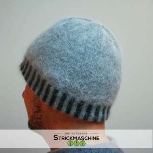 Rückenansicht eines Mannes mit Bart, der eine selbst gestrickte Beanie-Mütze aus flauschigem Angoragarn in hellem Eisblau mit dunkelgrau-gestreiftem Rippenbündchen trägt. Die Mütze hat eine weiche, zarte Textur. Darunter ein grauer Pullover. Vor neutralem hellgrauem Hintergrund. Unten das Logo von „Die Gläserne Strickmaschine".