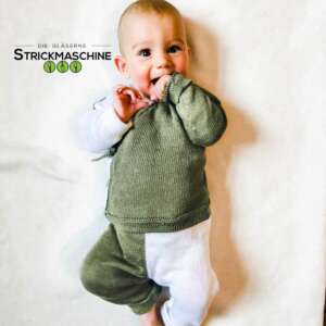 Ein fröhlich lächelndes Baby liegt auf dem Rücken und trägt ein selbst gestricktes zweiteiliges Set in Olivgrün und Weiß — bestehend aus einem Pullover mit weißen Ärmelbündchen und einer olivgrünen Hose mit weißem Einsatz. Das Baby steckt die Hand in den Mund und schaut aufgeweckt in die Kamera. Oben links das Logo von „Die Gläserne Strickmaschine".
