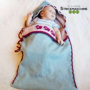 Ein Neugeborenes liegt wach und entspannt in einem selbst gestrickten Babyschlafsack in Hellblau mit bordeauxroten Zierkanten und Schnüren. Der Schlafsack hat eine Kapuze mit Wellenkante und einen rosafarbenen Innenkragen mit gestricktem Schaf-Jacquardmuster in Bordeaux. Das Baby trägt ein gestreiftes Kurzarm-Body. Oben rechts das Logo von „Die Gläserne Strickmaschine".