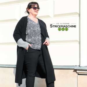 Eine Frau mit dunklem Haar und Sonnenbrille trägt einen selbst gestrickten langen schwarzen Cardigan mit grauen Strickbündchen an den Ärmeln, kombiniert mit einer schwarz-weiß gemusterten Bluse und schwarzer Hose. Sie steht vor einer hellen Hausfassade im Sonnenlicht. Oben rechts das Logo von „Die Gläserne Strickmaschine".