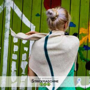 Rückenansicht einer Frau mit hochgestecktem, grau-lilafarbenem Haar, die einen selbst gestrickten Dreiecksschal in Naturbeige mit einem vertikalen petrolgrünen Zierstreifen als Schultertuch trägt. Darunter ein türkis-gemustertes Kleid. Sie steht vor einer bunt bemalten grünen Holzwand. Unten das Logo von „Die Gläserne Strickmaschine".