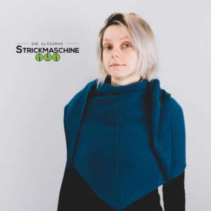 Eine Frau mit kurzem, grau-blondem Haar trägt einen selbst gestrickten großen Dreiecksschal in sattem Petrolblau, der als Schultertuch eng um Schultern und Brust gewickelt ist und eine markante dreieckige Spitze nach unten bildet. Darunter ein schwarzes Langarmshirt. Vor hellem, neutralem Hintergrund. Oben links das Logo von „Die Gläserne Strickmaschine".