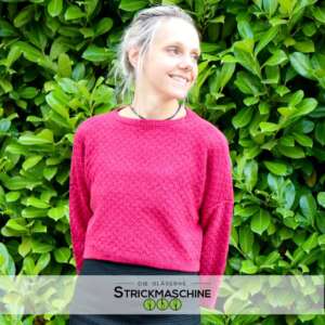 Eine lächelnde Frau mit hochgestecktem, grau-blondem Haar trägt einen selbst gestrickten kurzen Pullover in kräftigem Pink mit feinem Lochmuster und Rundhalsausschnitt. Dazu eine schwarze Hose. Sie steht vor einer dichten grünen Hecke und schaut seitlich nach oben. Unten das Logo von „Die Gläserne Strickmaschine".