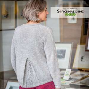 Rückenansicht einer Frau mit silbergrauem Bob, die einen selbst gestrickten hellgrauen Raglan-Pullover mit Zopfmuster-Details an den Schulternähten und einem dekorativen Schlitz mit Zopfmuster am unteren Rücken trägt. Dazu ein karierter Hosenrock in Pink-Rot. Im Hintergrund ein Innenraum mit gerahmten Kunstwerken. Oben rechts das Logo von „Die Gläserne Strickmaschine".