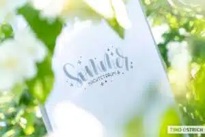 sommernachtstraum - Webinare und Kurse Weißes Papier mit dem Lettering „Sommer Nachtstraum" in Silber-Brushlettering und blauen Sternchen, fotografiert im Gegenlicht umgeben von unscharfen grünen Blättern und weißen Blüten. Foto: Timo Ostrich.