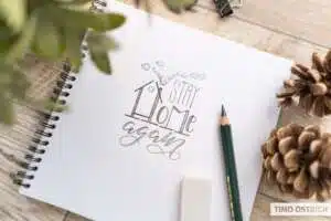 stay-home-again - Webinare und Kurse Spiralskizzenbuch auf einem Holztisch mit einer Bleistift-Lettering-Skizze: „Stay Home again" in gemischten Schriftstilen mit einem gezeichneten Haus und Rauchfahne. Daneben ein Faber-Castell-Bleistift, Radiergummi und zwei Tannenzapfen. Foto: Timo Ostrich.