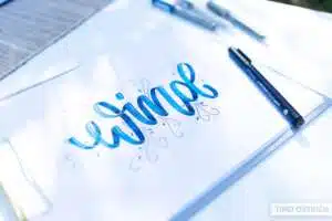 wind - Webinare und Kurse Handlettering „Wind" in kräftigem Blau mit dreidimensional wirkenden Buchstaben und zarten Schnörkellinien auf weißem Papier, eingeklemmt auf einem Klemmbrett im Freien. Daneben ein Tombow Mono Drawing Pen. Foto: Timo Ostrich.