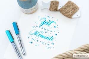 Handlettering „Zeit vergeht, Momente bleiben" in Türkis-Blau-Brushschrift mit Farbspritzern auf weißem Papier. Daneben zwei Brushmarker Pro in Türkis und Blau, ein Jutegarn auf einem Holzfischchen, ein Tau und ein rustikaler Blumentopf. Foto: Timo Ostrich.