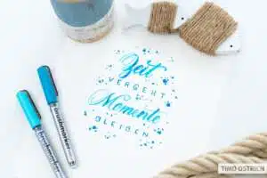zeit-vergeht-momente-bleiben - Webinare und Kurse Handlettering „Zeit vergeht, Momente bleiben" in Türkis-Blau-Brushschrift mit Farbspritzern auf weißem Papier. Daneben zwei Brushmarker Pro in Türkis und Blau, ein Jutegarn auf einem Holzfischchen, ein Tau und ein rustikaler Blumentopf. Foto: Timo Ostrich.