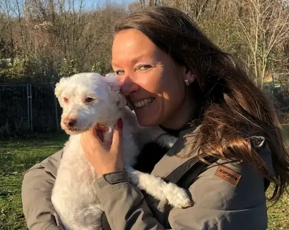 Lächelnde Frau mit langen braunen Haaren und grauer Jacke hält einen kleinen weißen Lockenhund (vermutlich Pudelmix) an ihre Wange gedrückt. Im Hintergrund eine grüne Außenanlage mit Bäumen und Zaun im Gegenlicht.