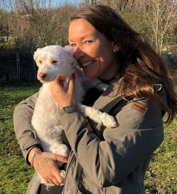 Lächelnde Frau mit langen braunen Haaren und grauer Jacke hält einen kleinen weißen Lockenhund (vermutlich Pudelmix) an ihre Wange gedrückt. Im Hintergrund eine grüne Außenanlage mit Bäumen und Zaun im Gegenlicht.