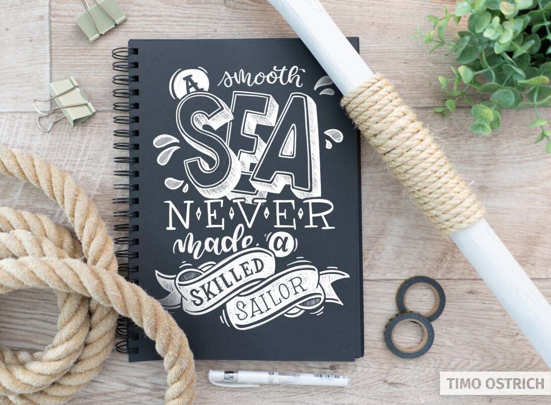 Kreideartiges Lettering „A smooth sea never made a skilled sailor“ auf einem schwarzen Notizbuch, arrangiert mit Seil und maritimen Elementen.