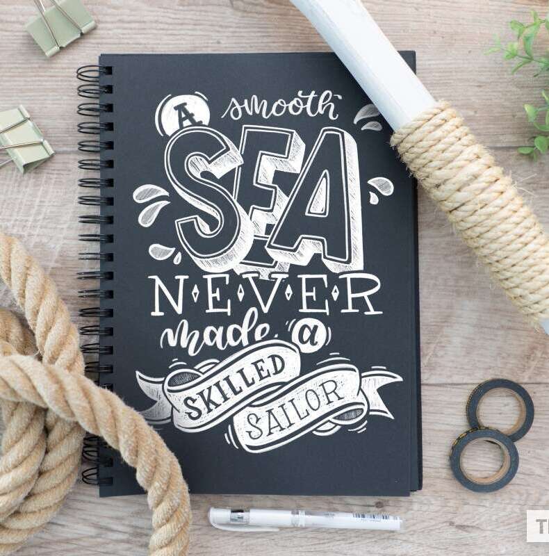 Kreideartiges Lettering „A smooth sea never made a skilled sailor“ auf einem schwarzen Notizbuch, arrangiert mit Seil und maritimen Elementen.