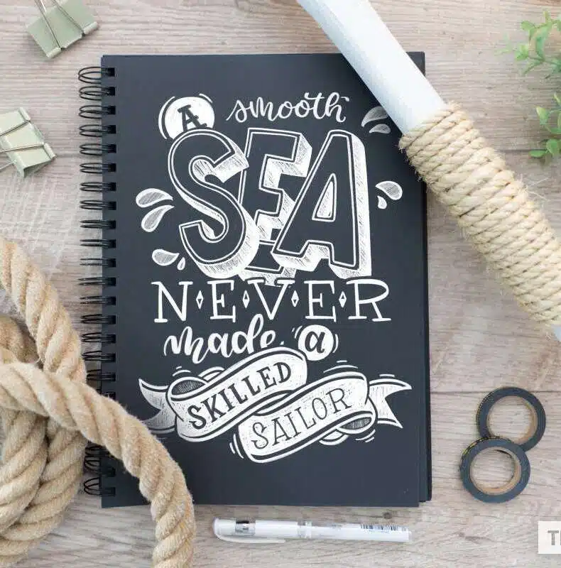 Kreideartiges Lettering „A smooth sea never made a skilled sailor“ auf einem schwarzen Notizbuch, arrangiert mit Seil und maritimen Elementen.