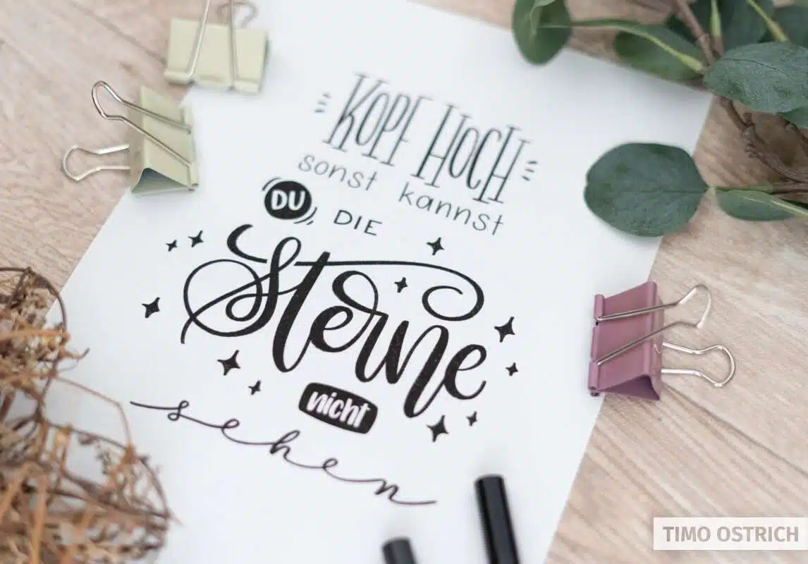 Handlettering „Kopf hoch, sonst kannst du die Sterne nicht sehen“ auf Papier, dekoriert mit Sternen, Blättern und Klammern.