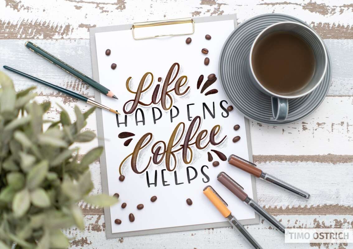 Handlettering „Life happens, coffee helps“ auf Papier neben einer Kaffeetasse, Stiften, Kaffeebohnen und Pflanze.