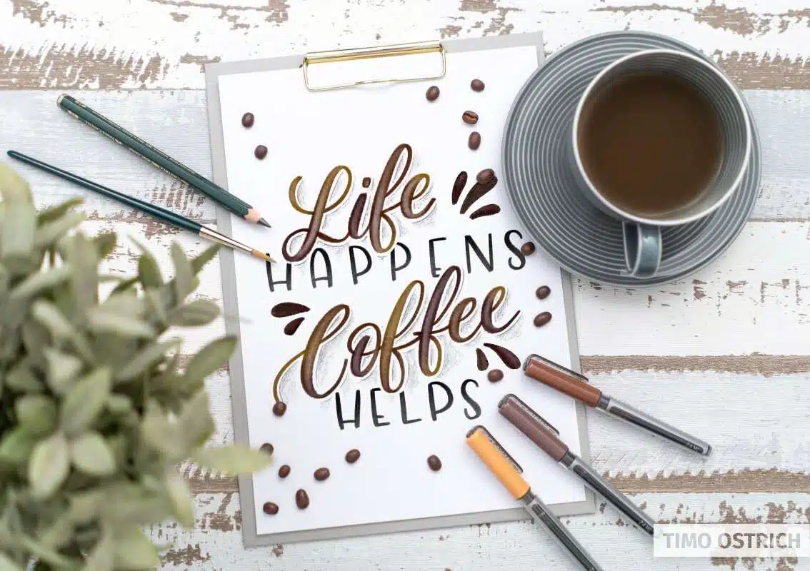 Handlettering „Life happens, coffee helps“ auf Papier neben einer Kaffeetasse, Stiften, Kaffeebohnen und Pflanze.