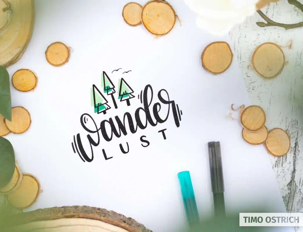 Handlettering mit dem Wort „Wanderlust“ auf weißem Papier, umgeben von Holzscheiben, Blättern und Stiften.