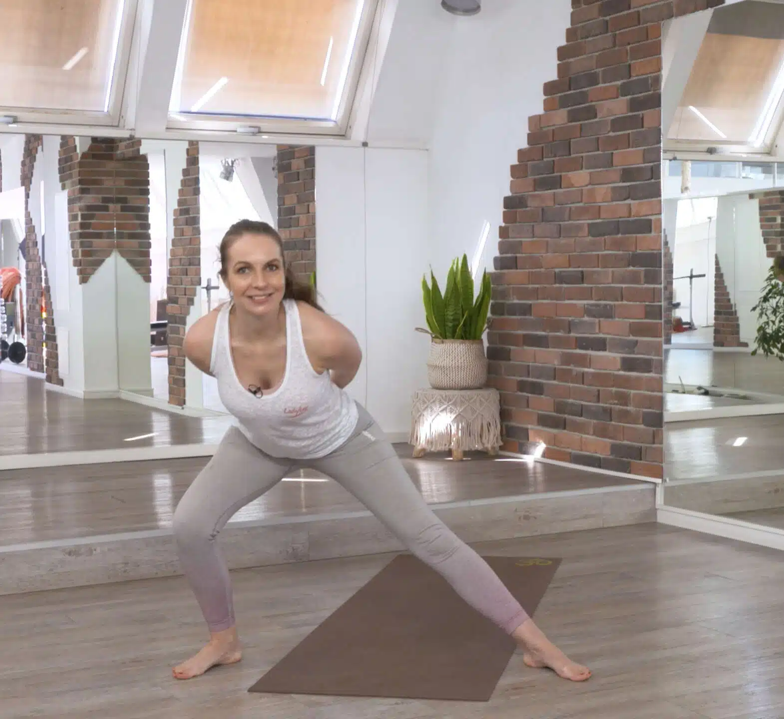 Screenshot eines Pilates-Onlinekurses: Eine Trainerin in weißem Top und hellgrauen Leggings steht in einer breiten Grätsche mit leicht gebeugten Knien vor einer braunen Matte. Im Hintergrund ein helles Studio mit Backsteinwänden, Spiegeln und Zimmerpflanzen.