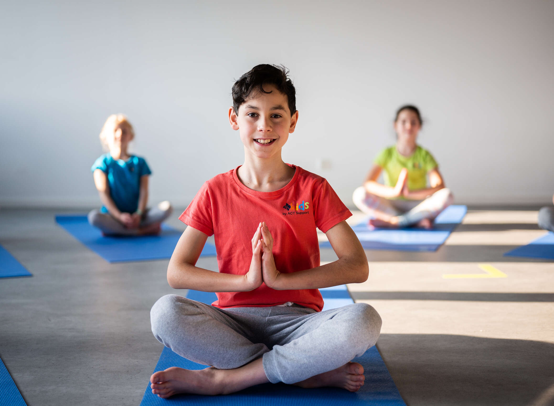 Lächelnder Junge im roten T-Shirt sitzt im Schneidersitz auf einer blauen Yogamatte mit Händen in Namaste-Haltung. Im Hintergrund weitere Kinder auf blauen Matten in einem hellen Yogaraum.