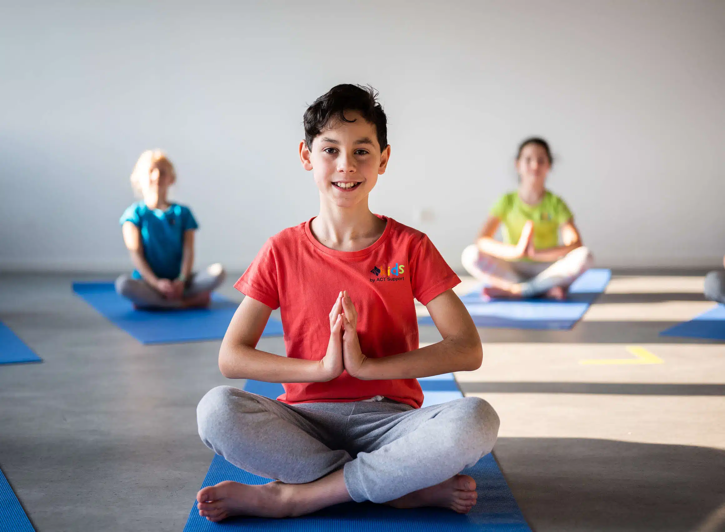 Lächelnder Junge im roten T-Shirt sitzt im Schneidersitz auf einer blauen Yogamatte mit Händen in Namaste-Haltung. Im Hintergrund weitere Kinder auf blauen Matten in einem hellen Yogaraum.
